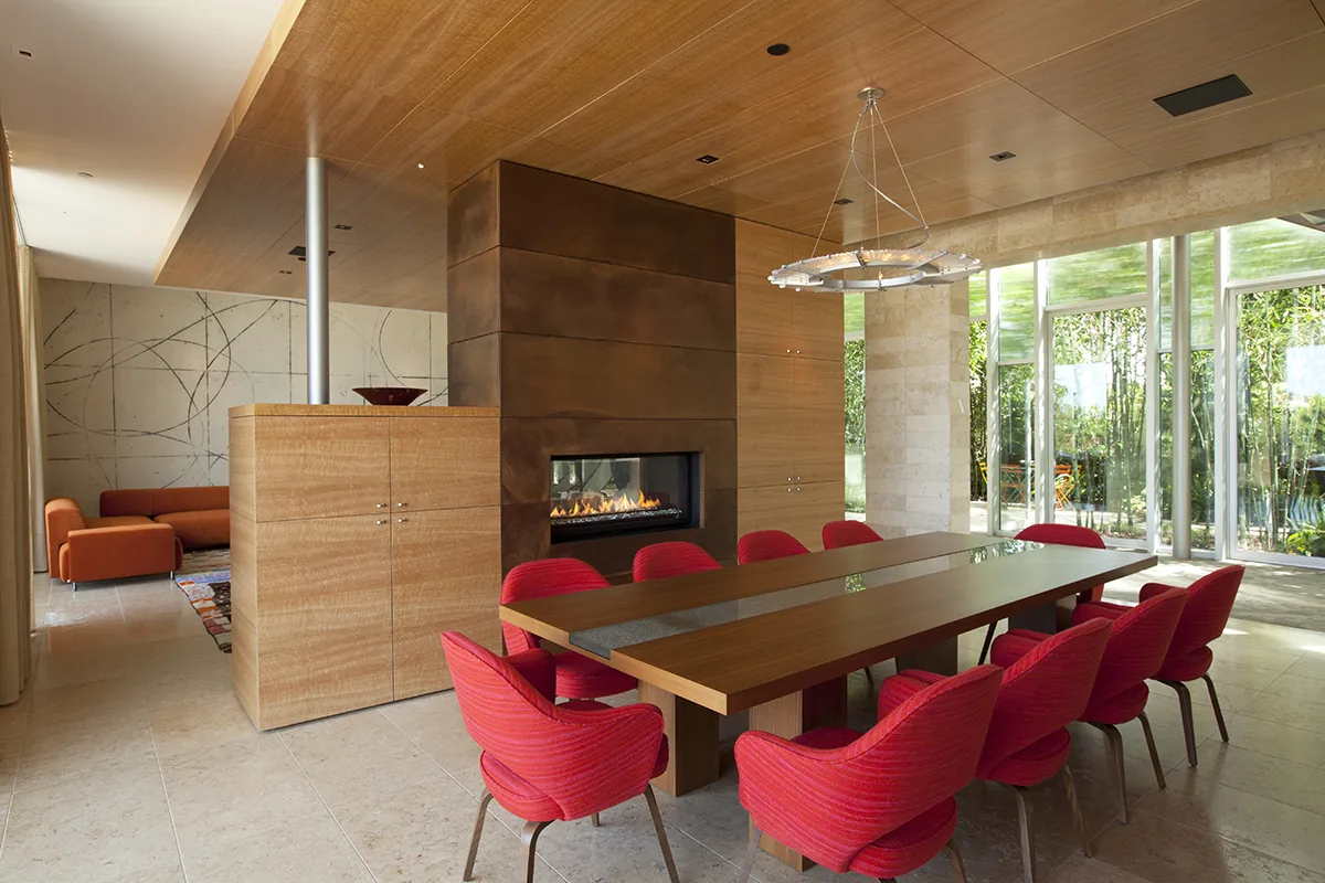 Dining area features a long wooden table, red chairs, and a built-in fireplace, illuminated by a circular chandelier.