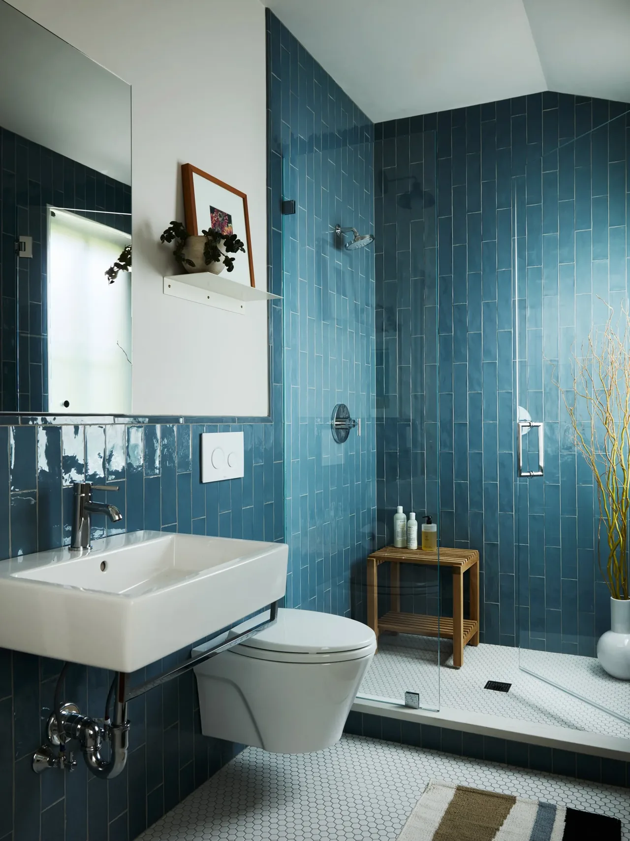 Bathroom with wall-mounted sink and toilet, blue tile walls, and walk-in shower with glass door and wooden bench.
