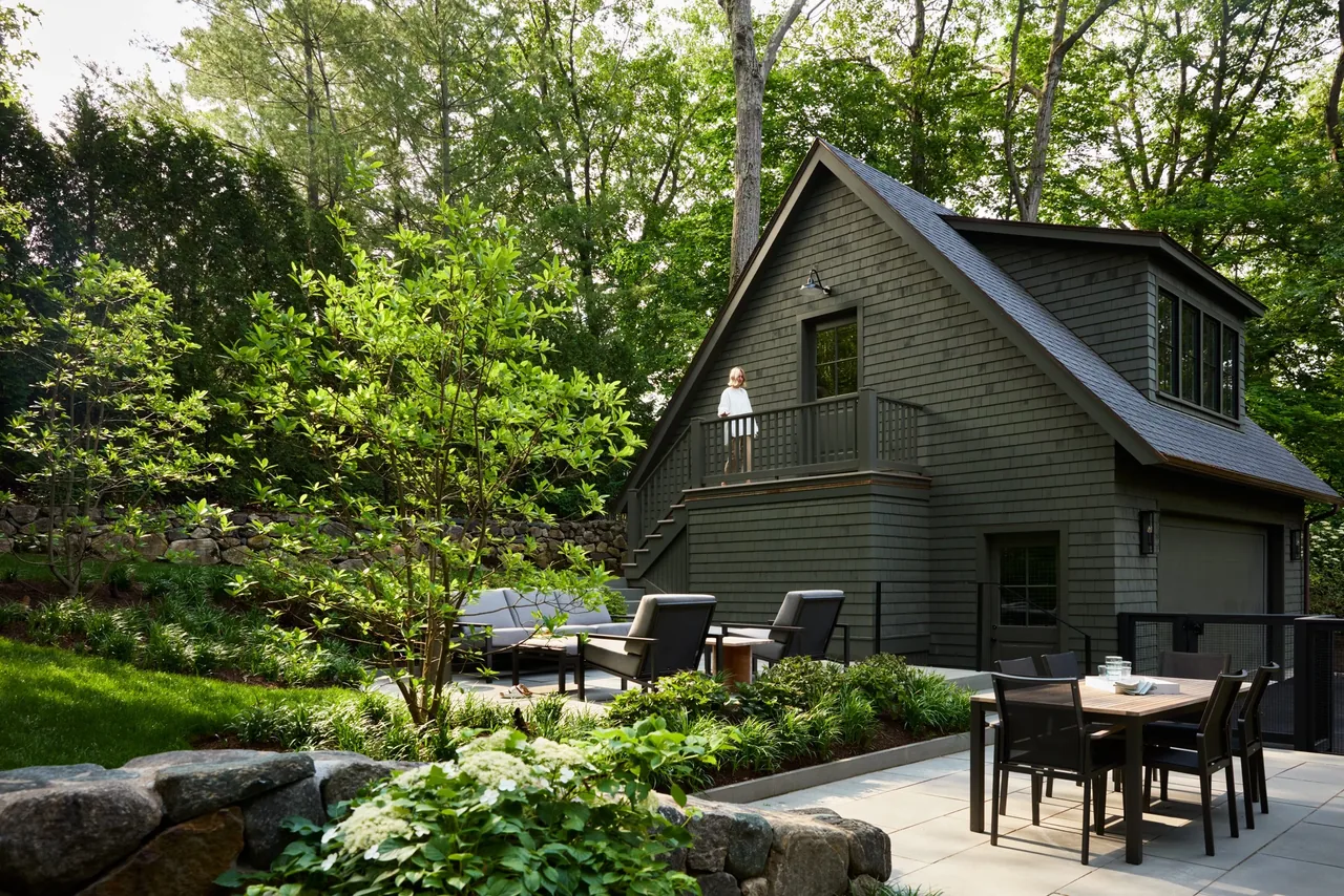 Patio and outdoor seating area next to a two-story shingle-sided building with dormer windows and entry stairs.