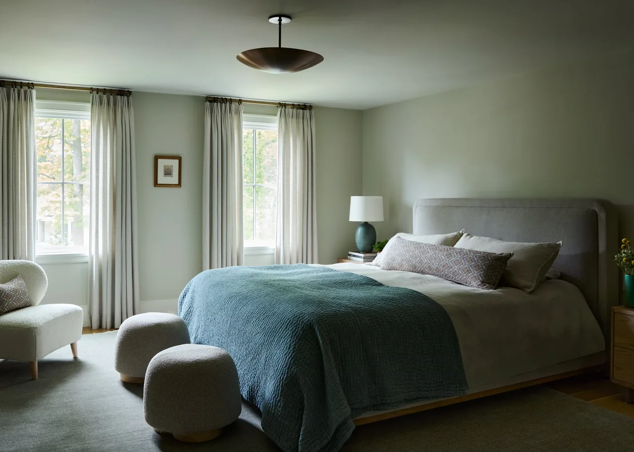 Bedroom featuring a light gray upholstered bed, blue quilt, neutral tones, and simple window drapery with a sitting chair and ottomans.