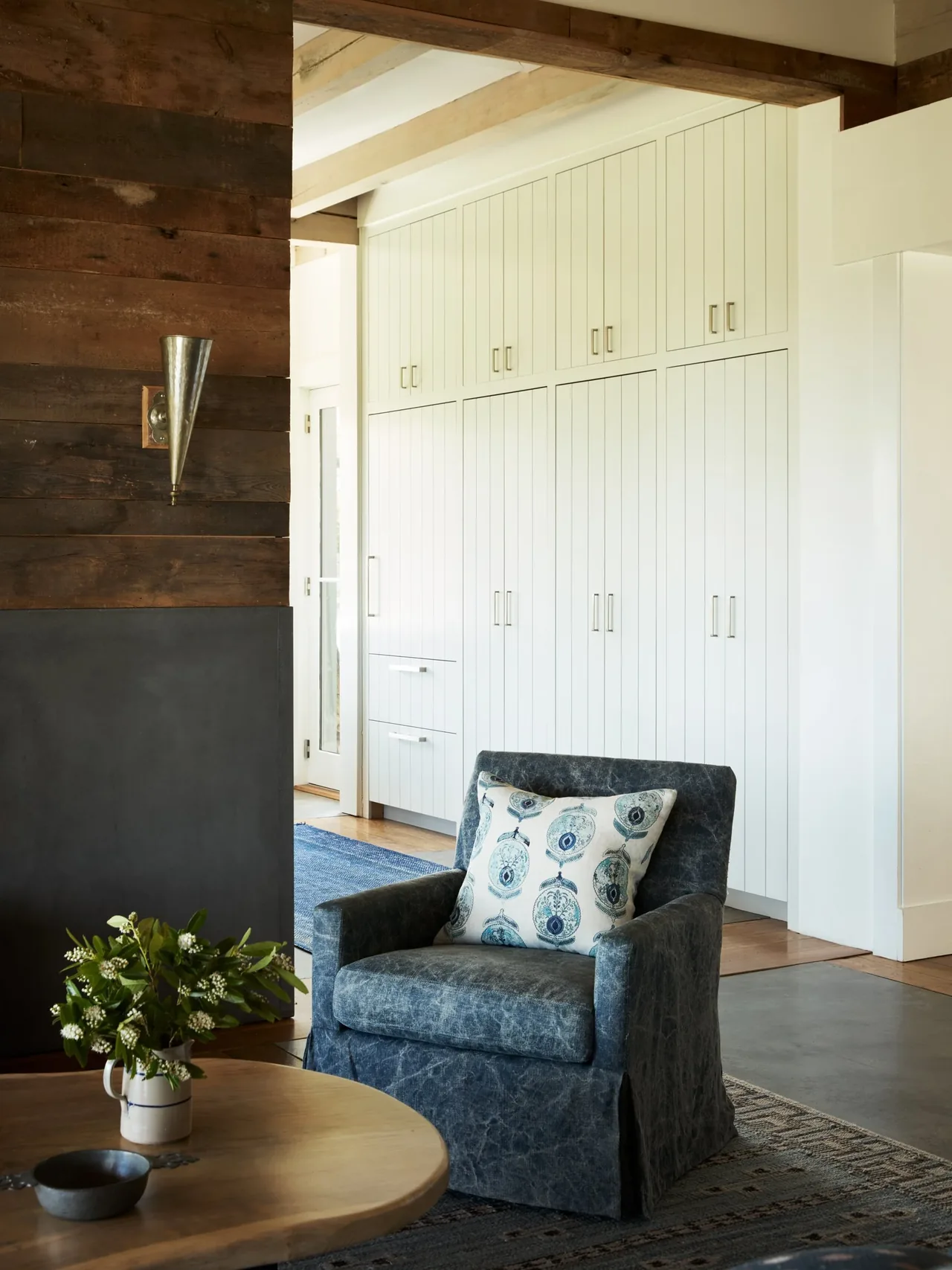 A seated area features a deep blue armchair and extensive white vertical paneled cabinetry under exposed wood beams.
