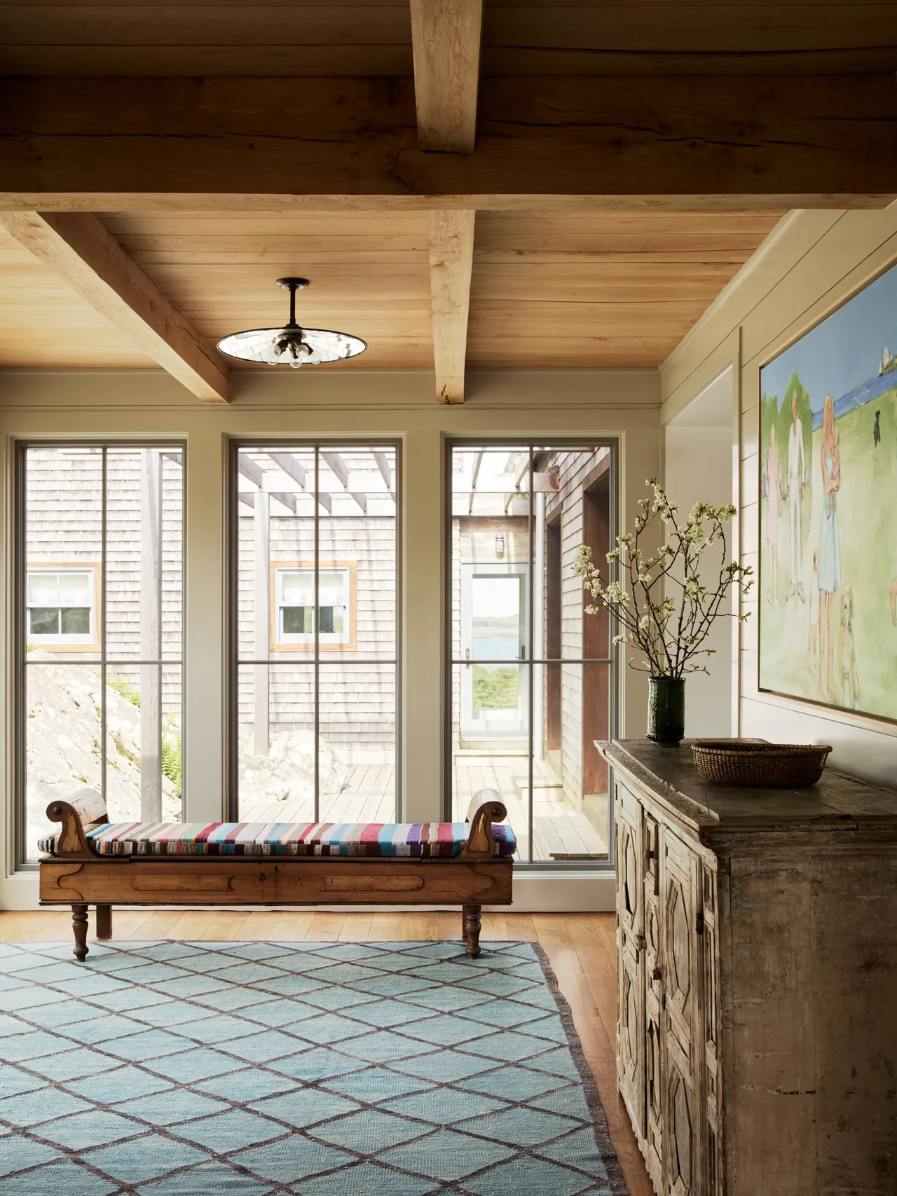 Room features wide wooden beams, large windows with multiple panes, a colorful fabric bench, and a rustic cabinet under a mounted painting.