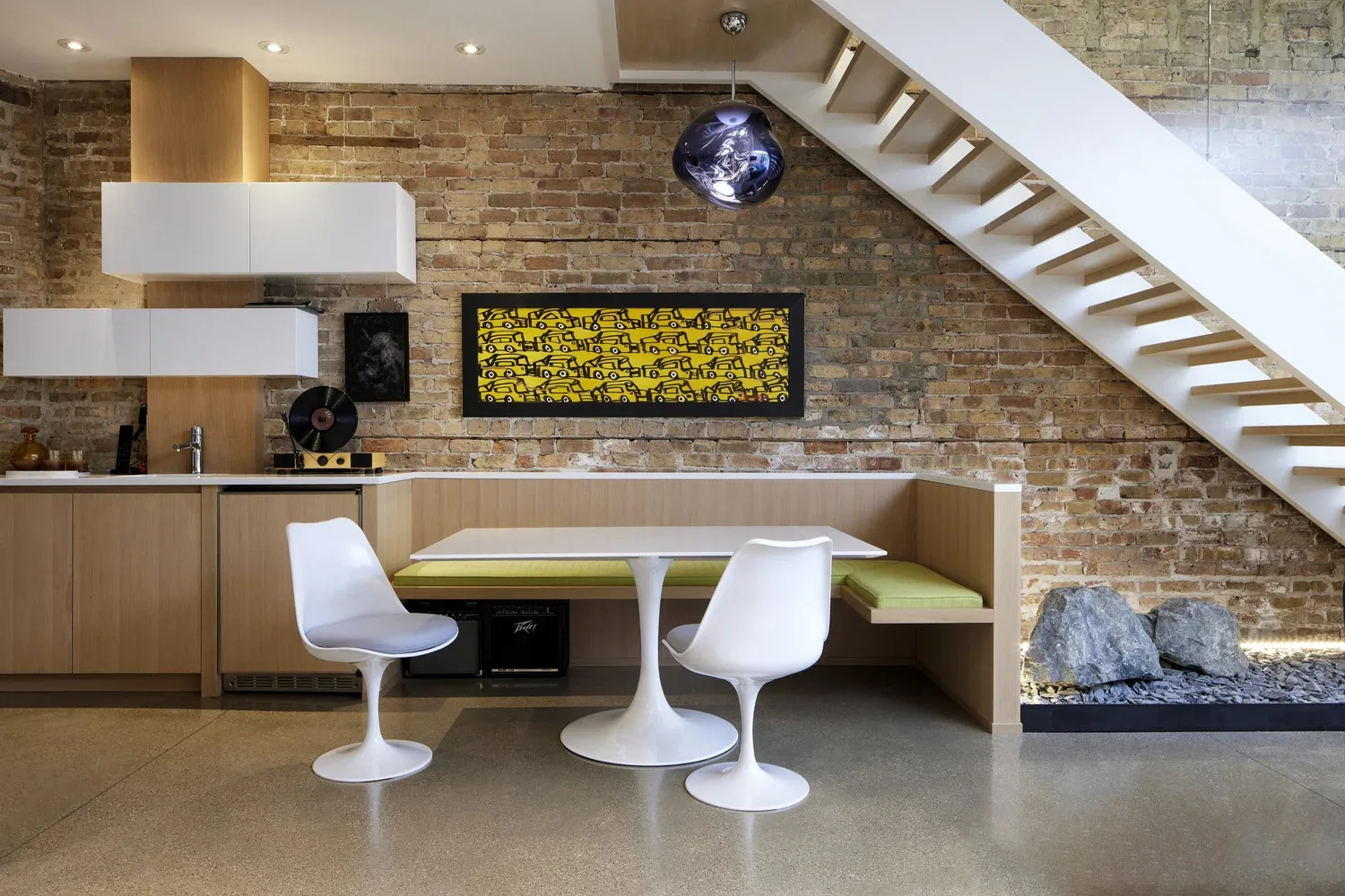 Kitchen and dining nook with exposed brick wall, tulip chairs, and built-in banquette under staircase.