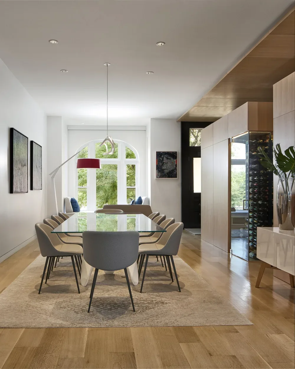 Dining room features a glass table, upholstered chairs, wooden flooring, and a large arched window with a view of greenery.