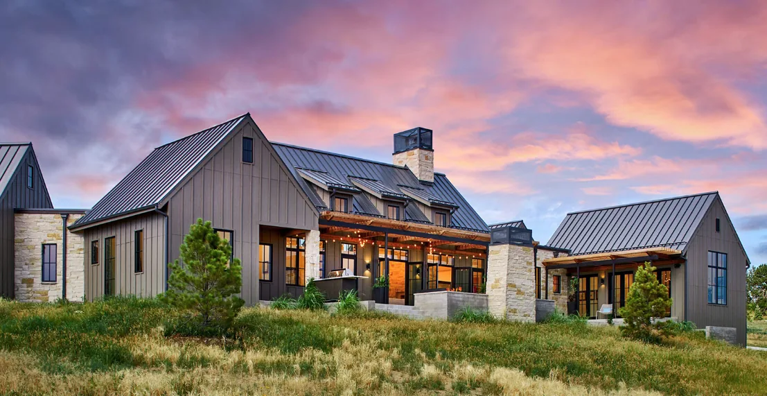 Contemporary farmhouse exterior featuring stone chimneys, standing seam metal roofs, and black window frames at twilight.
