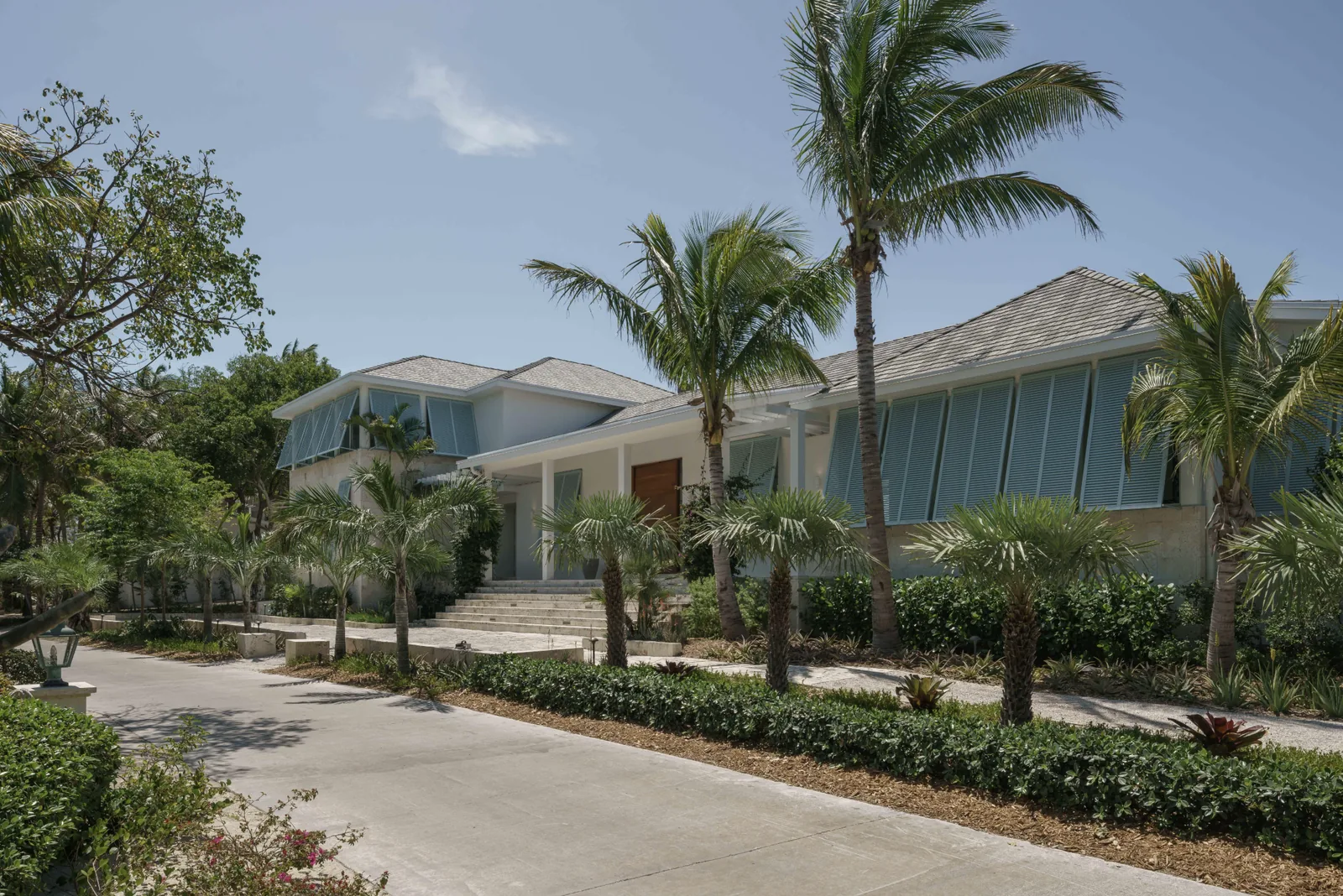 Exterior view of Cat Cay Residence showing a one-story building with a pitched roof, blue window shutters, and a landscaped front with palm,