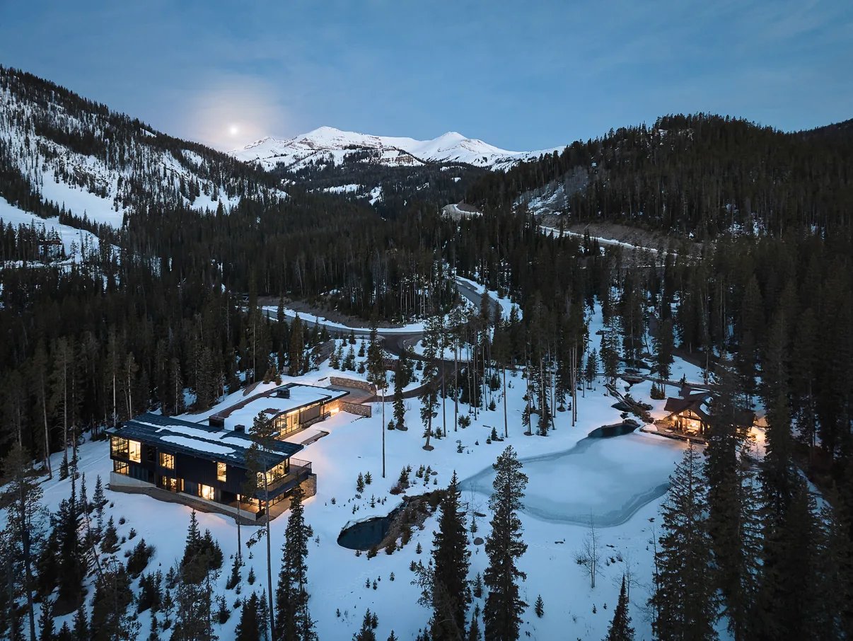Modern house sits in a snowy mountain forest with warm lights glowing inside and an icy pond nearby.