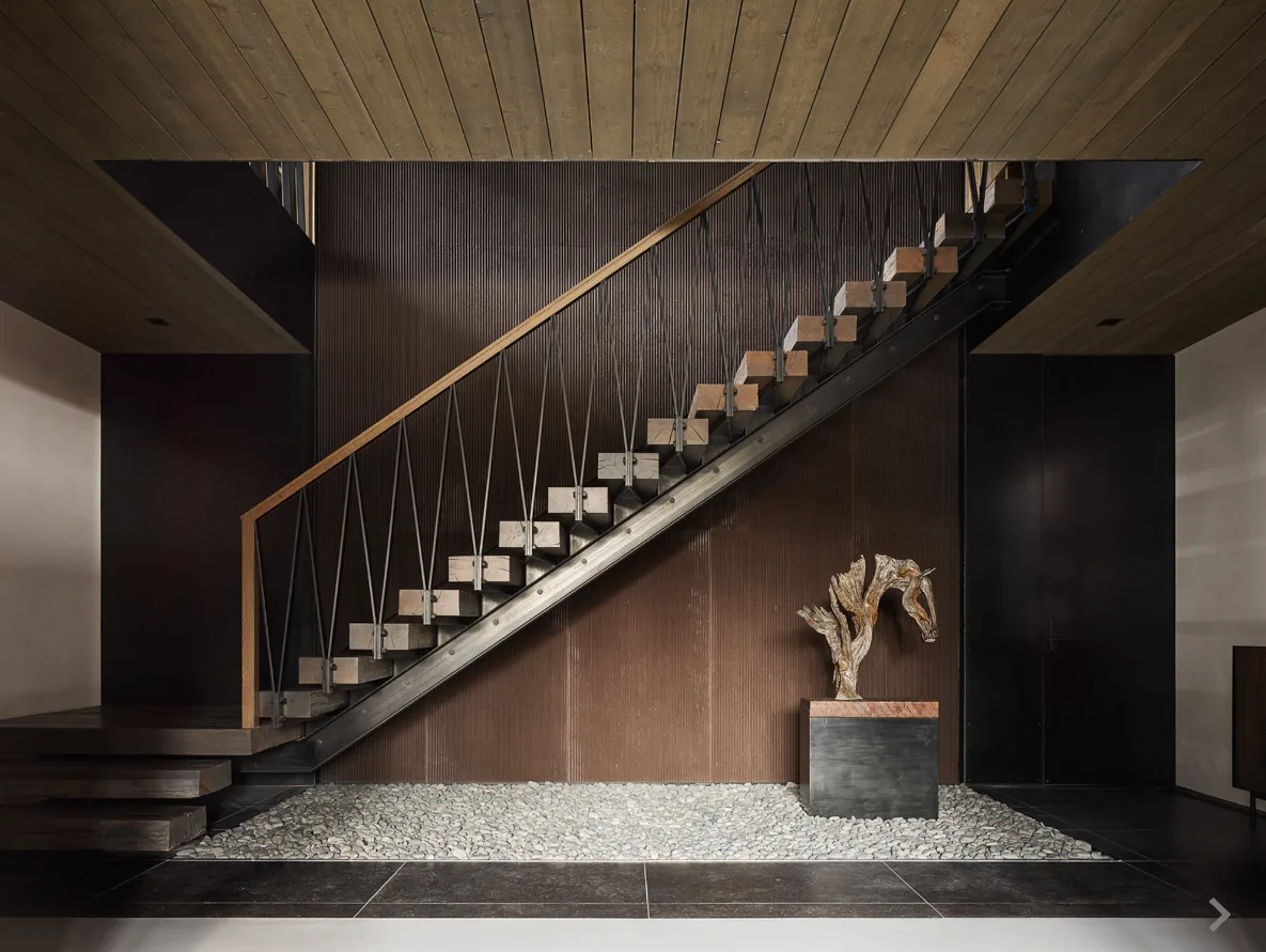 Modern stair with metal and wood materials above a pebble bed and a wooden sculpture on a pedestal.