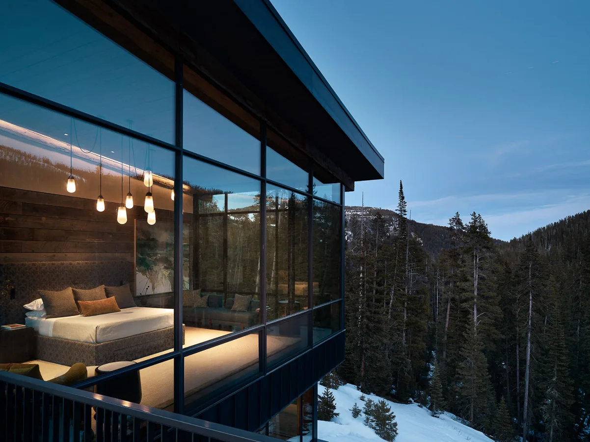 Bedroom with floor-to-ceiling glazing offers views of snowy pine forest and mountains at dusk.