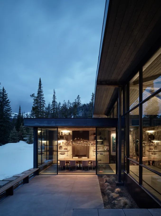 Modern dining room is visible through floor-to-ceiling black metal framed windows in a snowy forest setting.