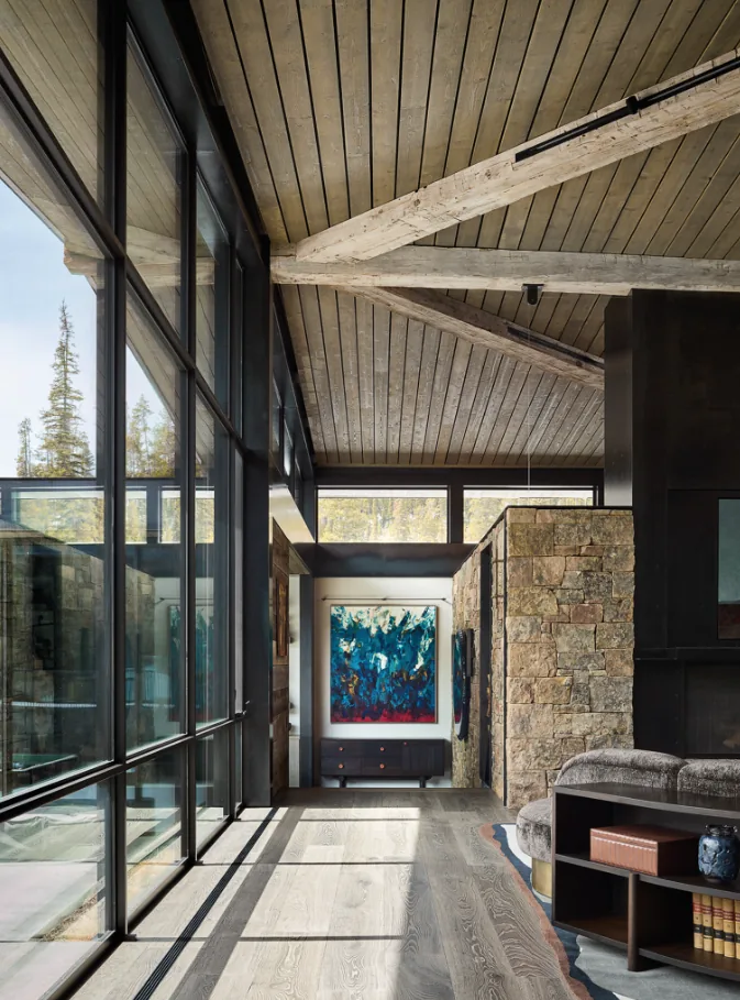Room features floor-to-ceiling glazing with black steel framing, a vaulted wood ceiling with exposed beams, and a stone wall interior.