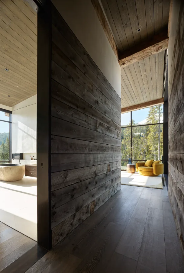A hallway with wood plank walls opens into a living space featuring floor-to-ceiling glazing with mountain views and a yellow chair.