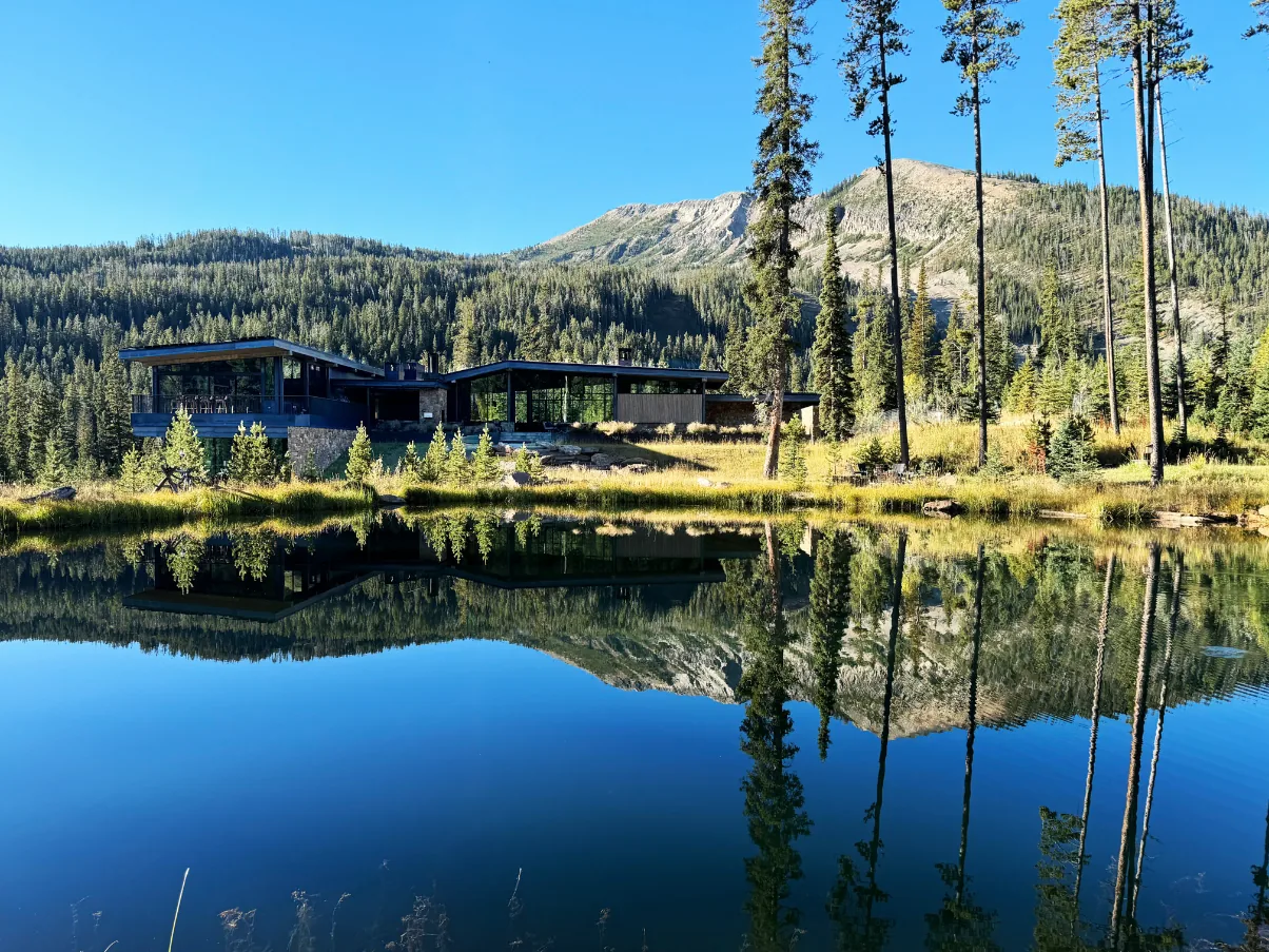 Modern house with large glass windows reflected in a calm pond, surrounded by pine trees and mountains.
