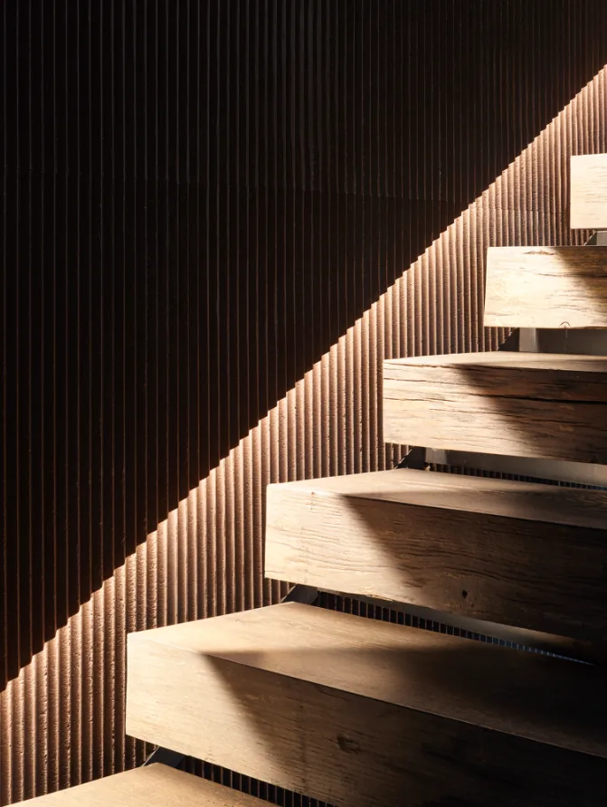 Floating wooden stairs illuminated by angled natural light on a vertically grooved wood wall.