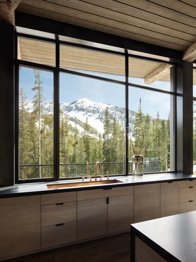 Kitchen sink area with black metal window frames showcasing a snowy mountain and forest panorama.