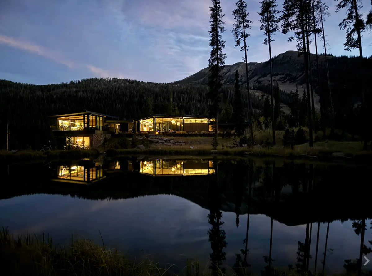 Night view of Yellowstone project home with expansive glass walls reflecting in lake, set against pine trees and mountains.