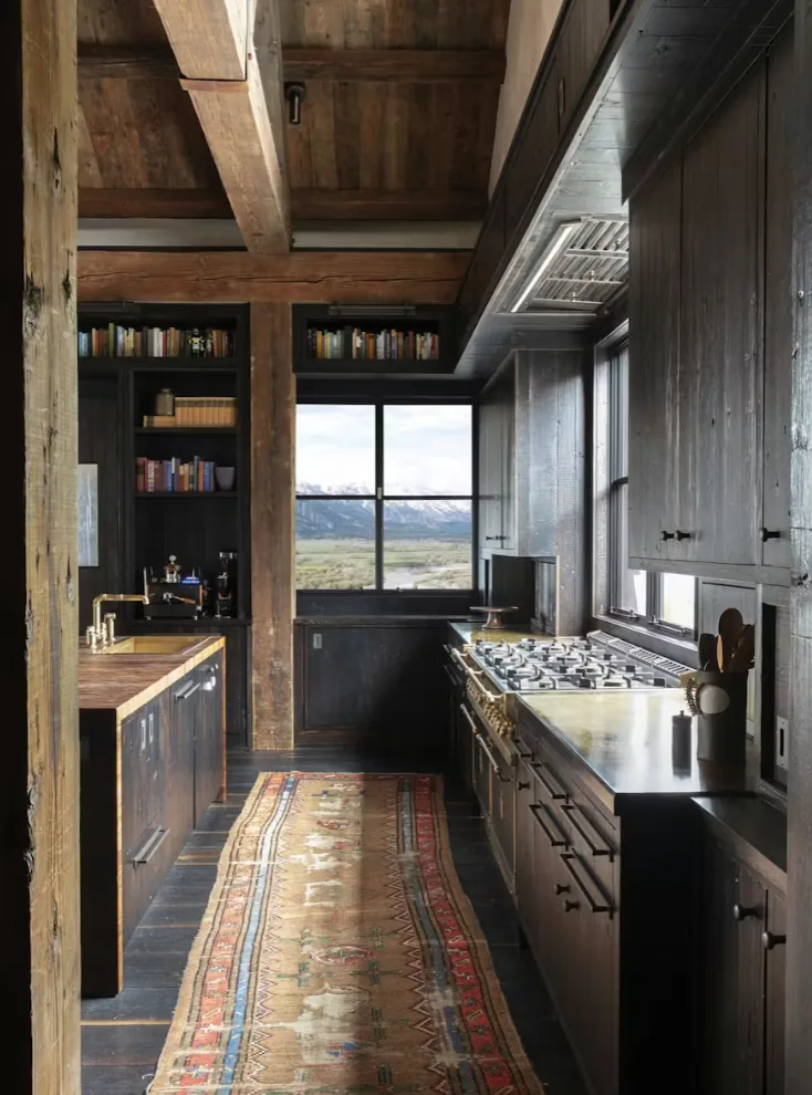 Kitchen with dark wood interiors and a view of snowy mountains through the window, featuring a gas range and rustic wooden ceiling beams.
