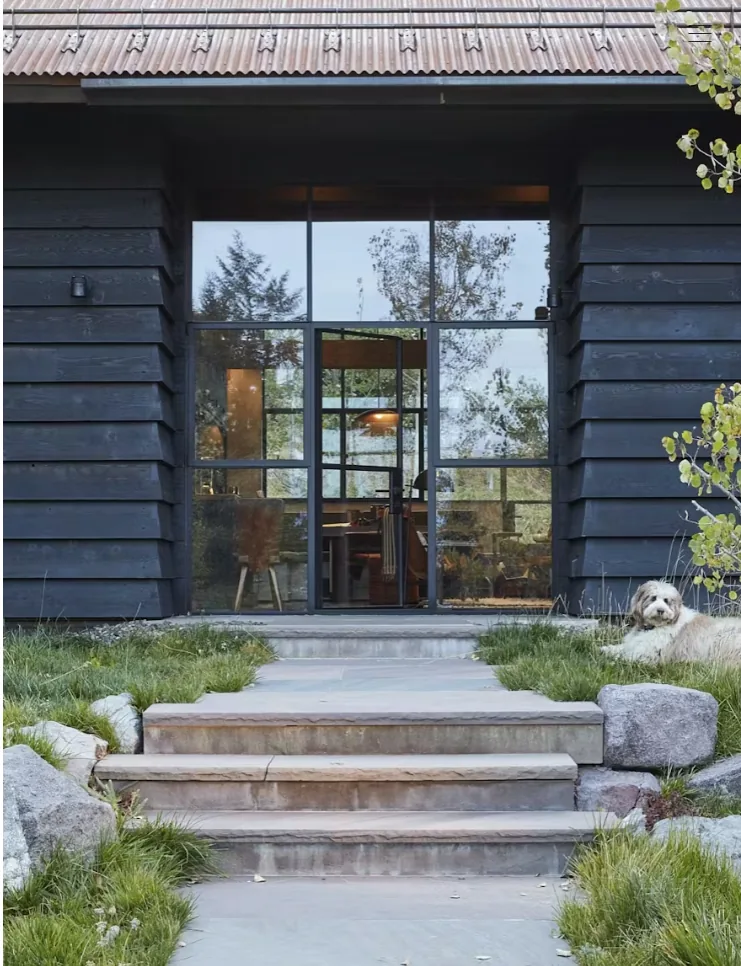 Camp Teton entry features black wood siding, rusted metal roof, and large black-framed glass door with visible interior lighting.