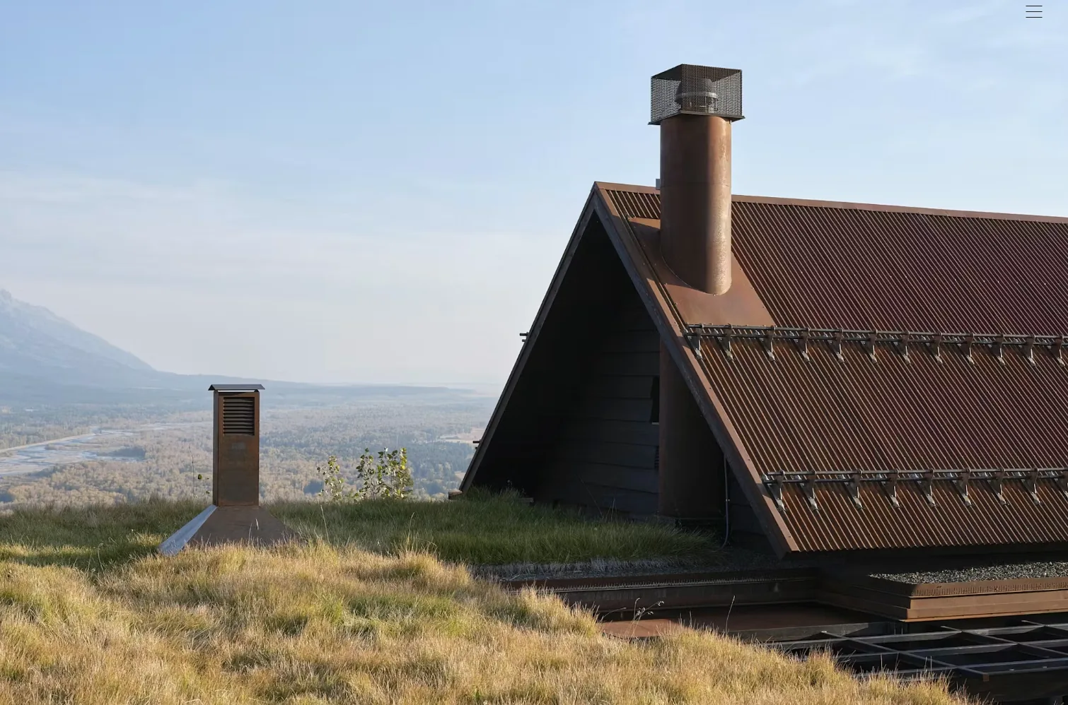 Camp Teton features a rusted metal gable roof with chimney set in a grassy hillside overlooking a mountain landscape.