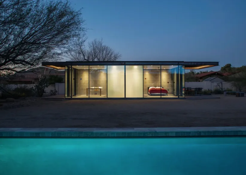 Night view of a single-story modern glass and concrete house with interior lights on and pool in front.