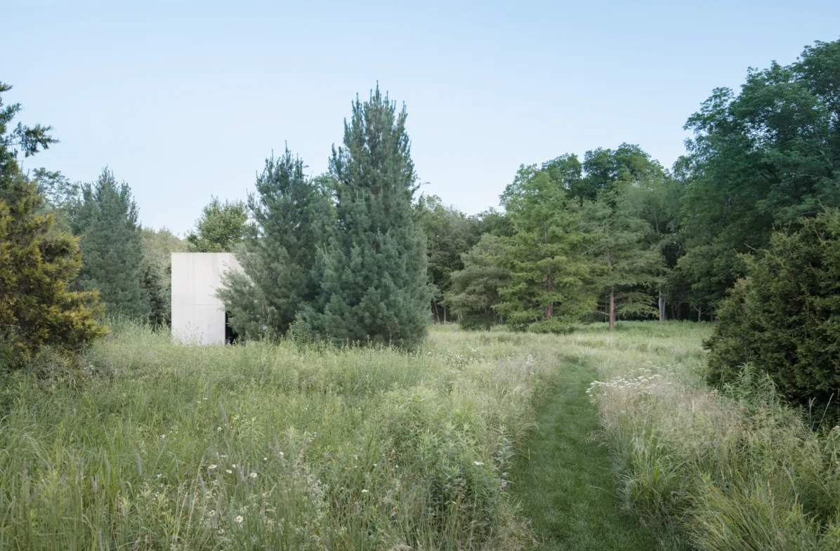 This image shows a green field with a narrow path leading to a small concrete building, surrounded by various trees and vegetation.