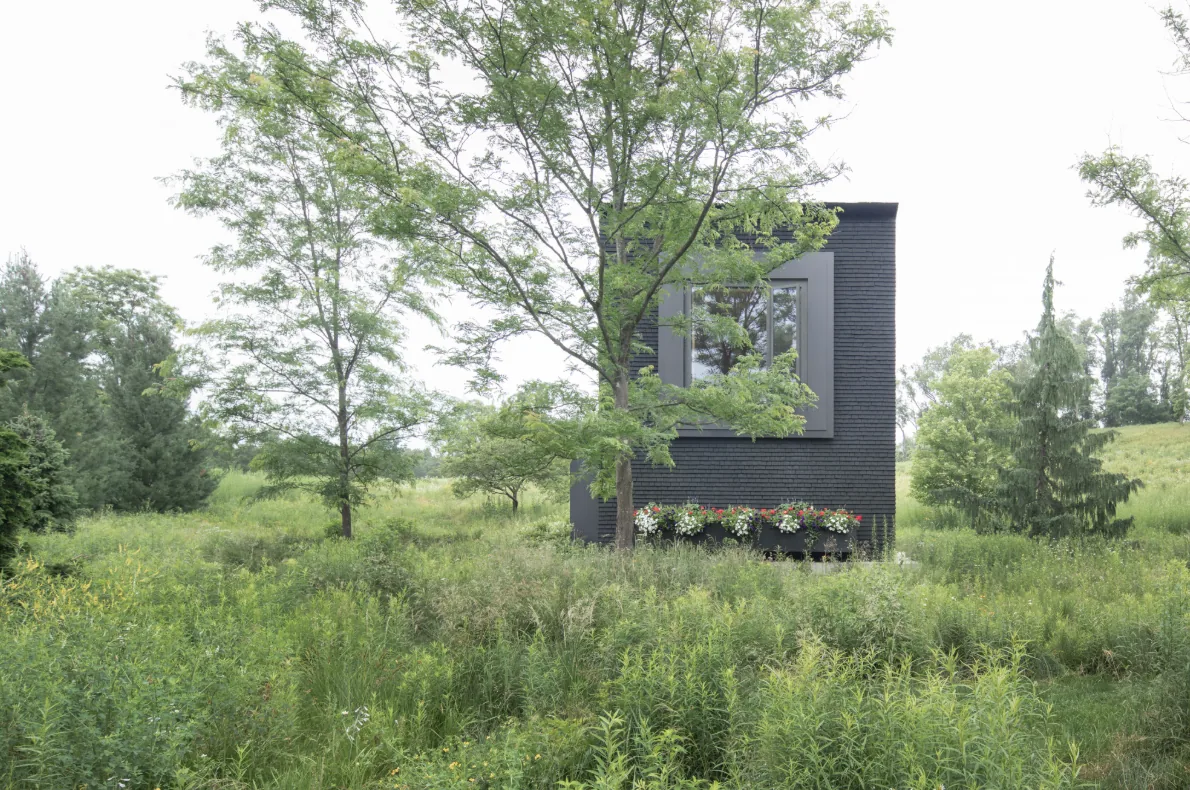 Hudson Valley House II features a dark exterior with large recessed window and lush surrounding vegetation.