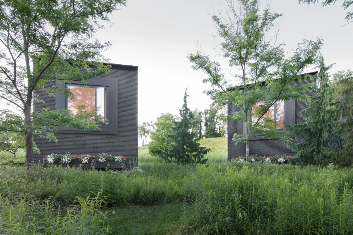 Exterior view of two black brick houses with large recessed windows and surrounding dense planting in a rural setting.