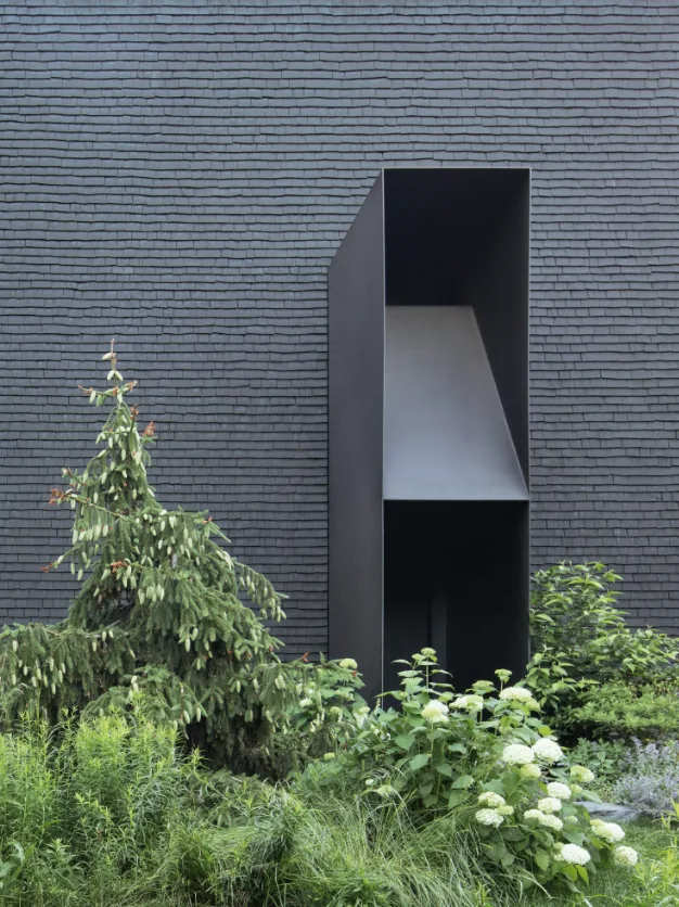 Hudson Valley House II features dark shingle siding with a large black recessed window opening and dense surrounding plantings.