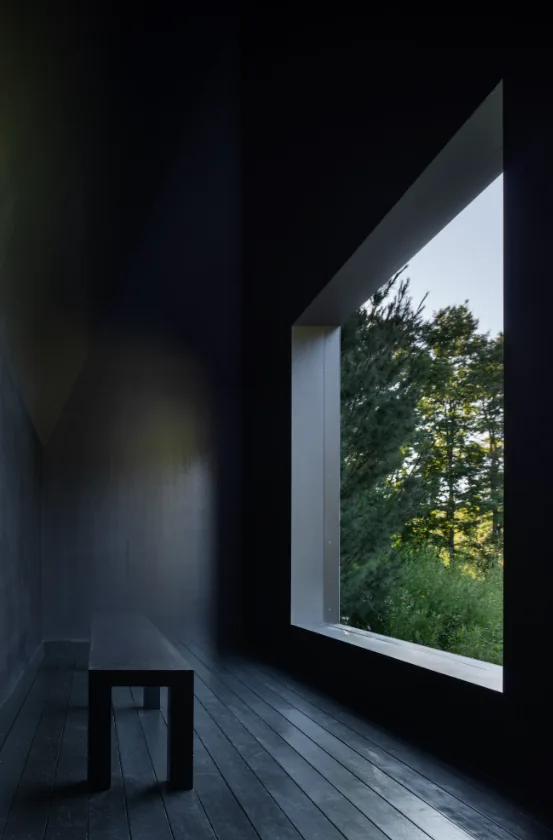 Interior view of a dark wood-floored room with a large window framing trees and greenery in Hudson Valley House II.