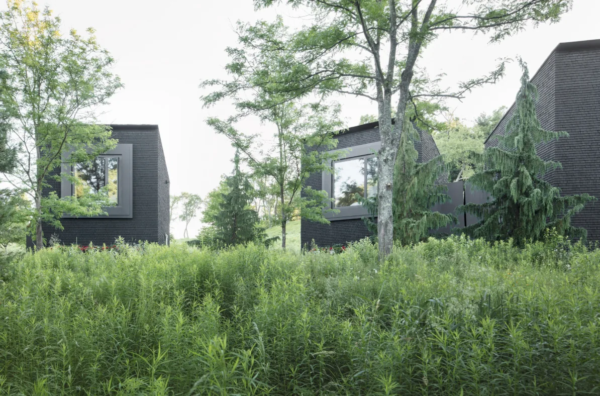 Hudson Valley House II features dark shingle siding and large window openings amidst natural greenery.