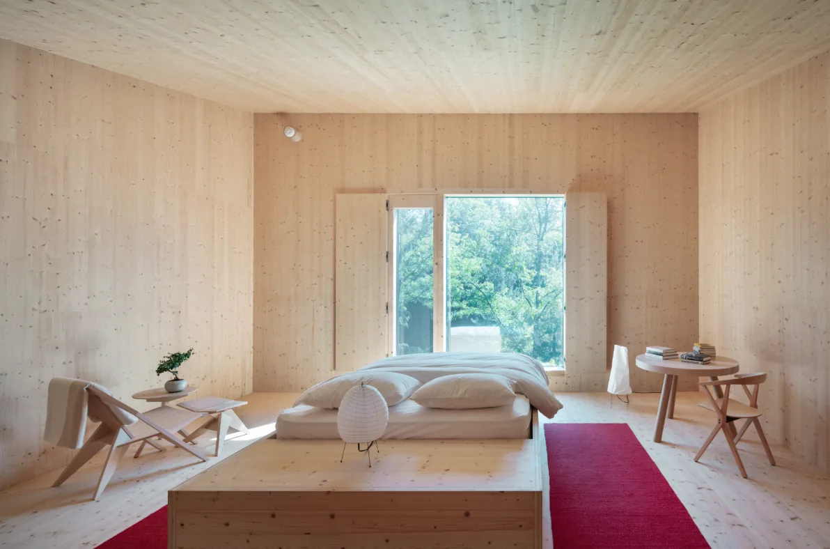 Bedroom with light wood panel walls and ceiling, floor-to-ceiling window revealing lush greenery outside.