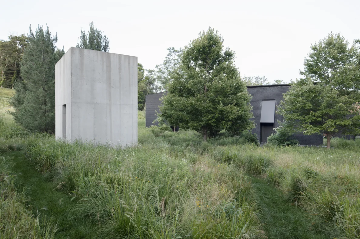 Concrete cubical structure near a dark brick house with large windows in a lush, grassy landscape.