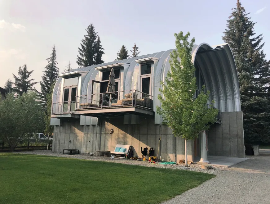 A modern house with a curved metal roof and concrete base surrounded by trees and green lawn.