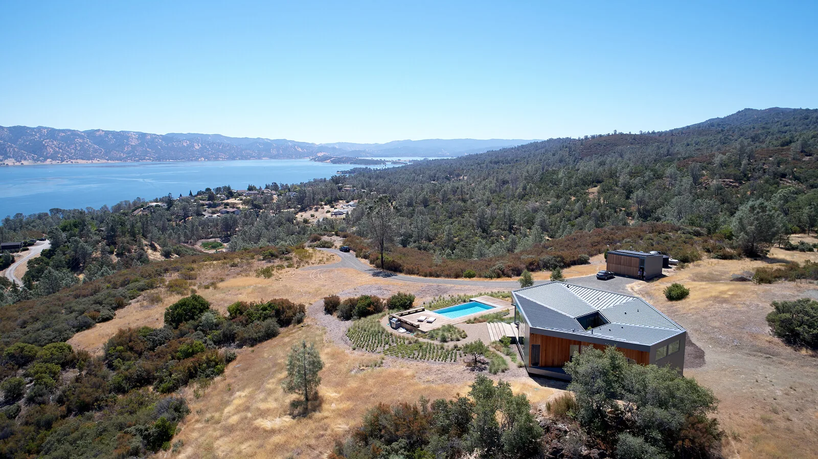 A modern house with a metal roof and wooden siding on a hillside overlooking a large body of water and forested mountains.