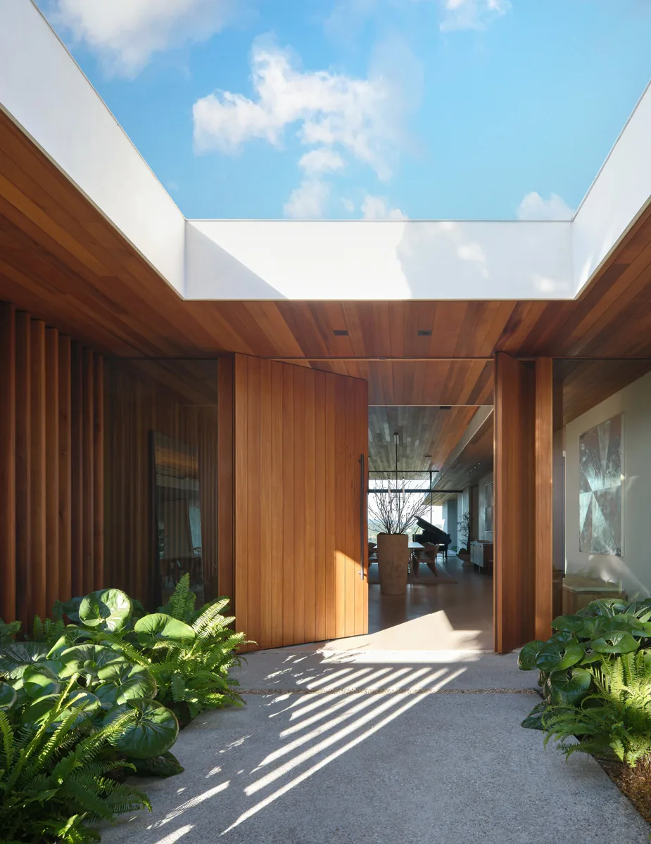 The Carla Ridge House entry features a large wooden pivot door with floor-to-ceiling glass walls and lush greenery under a skylight.
