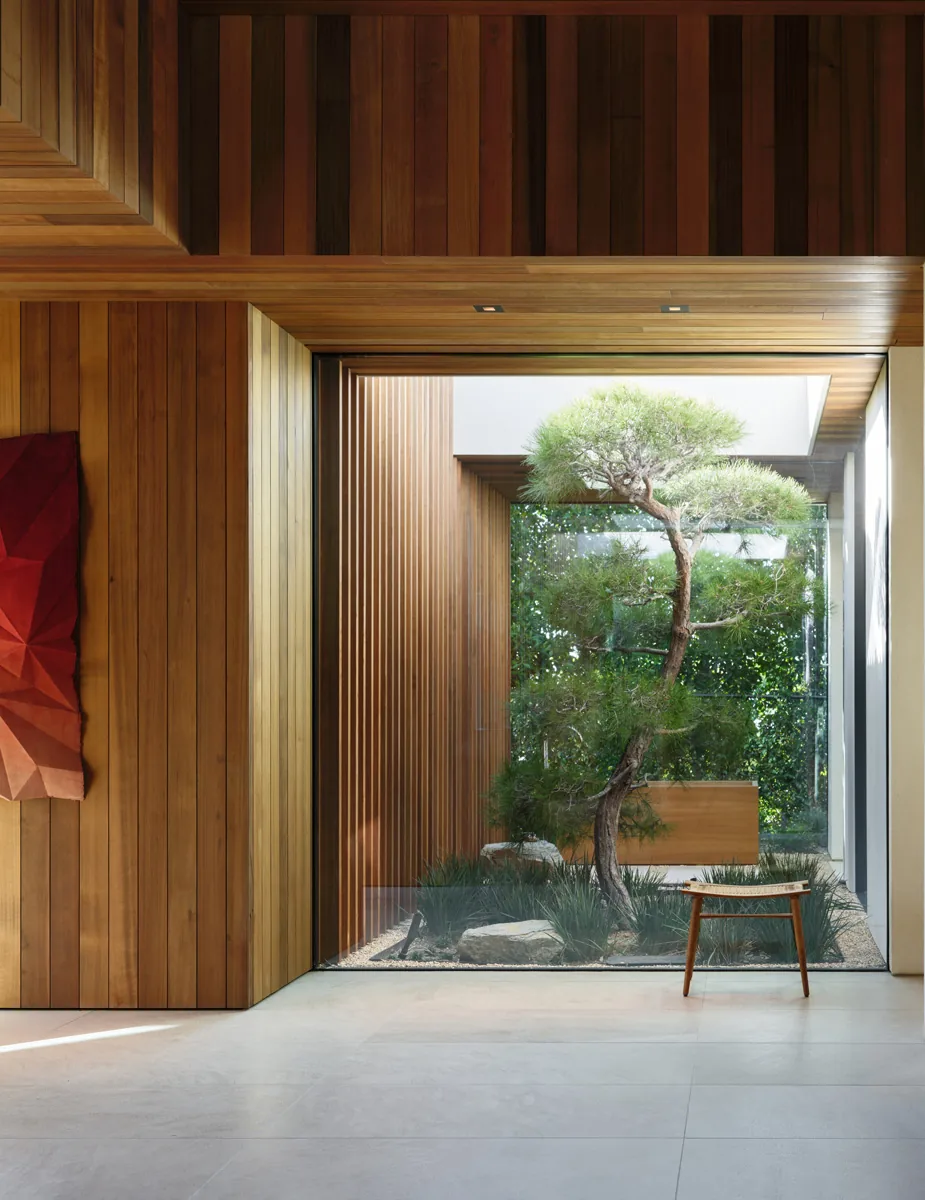 The Carla Ridge House features a wood-paneled interior wall and ceiling with a large glass window revealing an indoor courtyard with natural landscaping and sculpted tree.