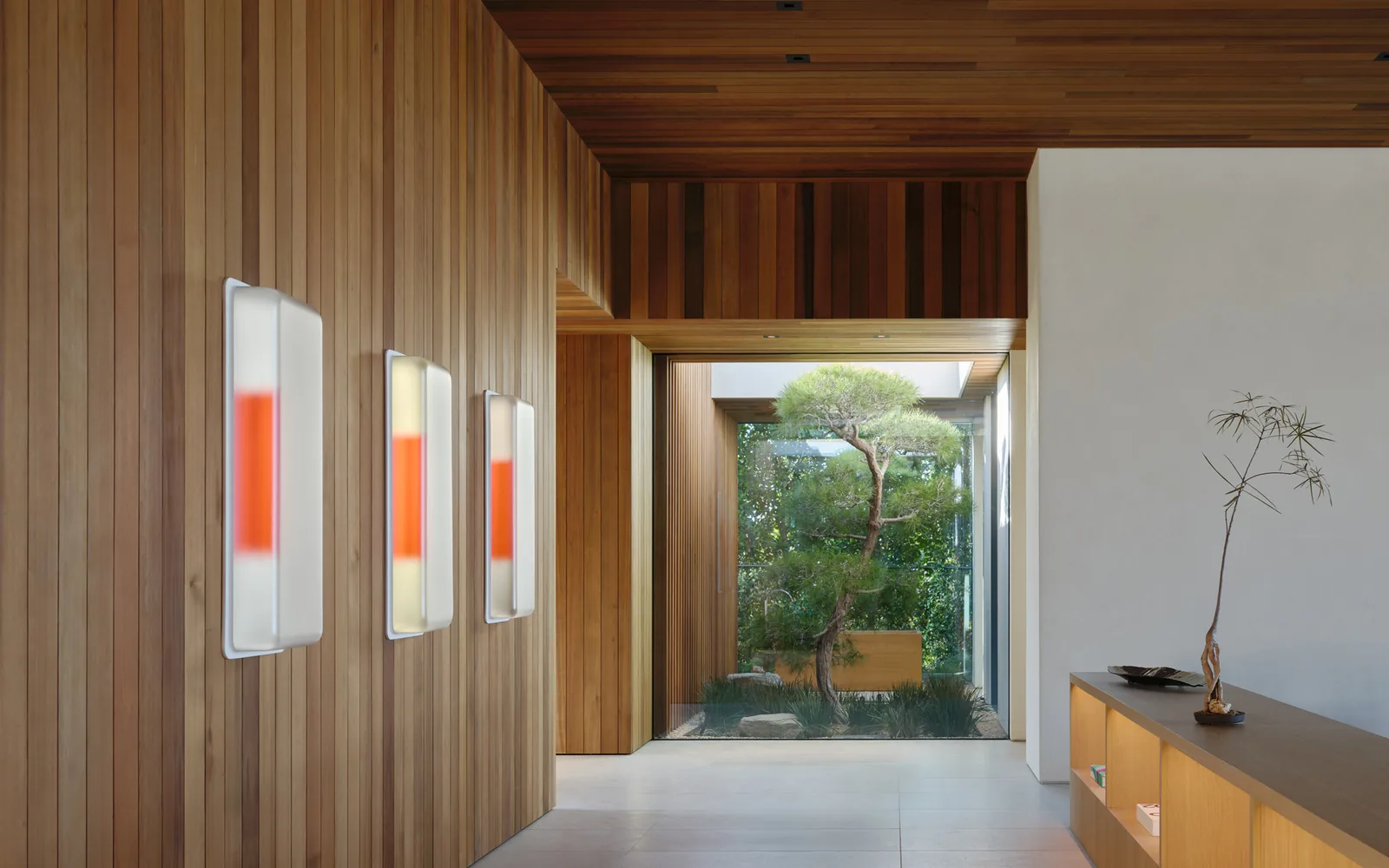 Wood-paneled hallway with three modern wall sconces and a view into a glass-enclosed courtyard with a tree at Carla Ridge House.