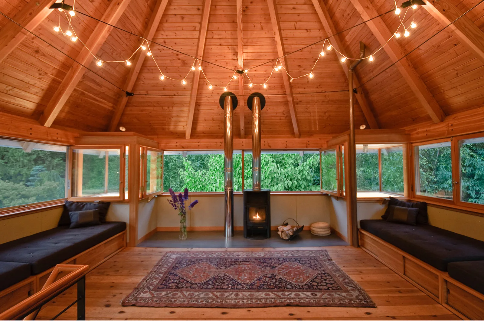 Wood-paneled interior room features black cushioned benches, a wood stove with dual metal chimneys, and string lights under a peaked ceiling.