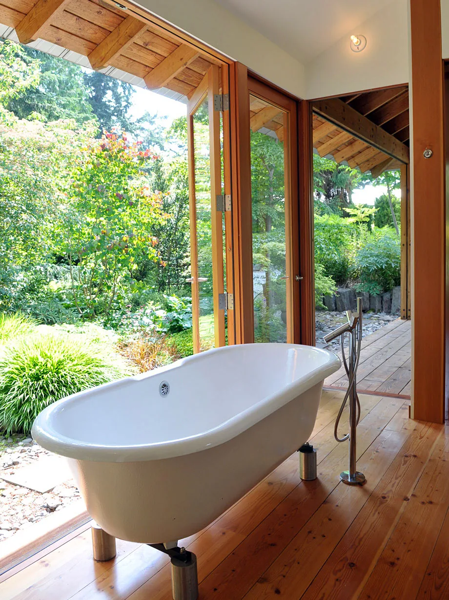 Bathroom with freestanding tub opening onto a wooden deck and lush garden through large glass doors.