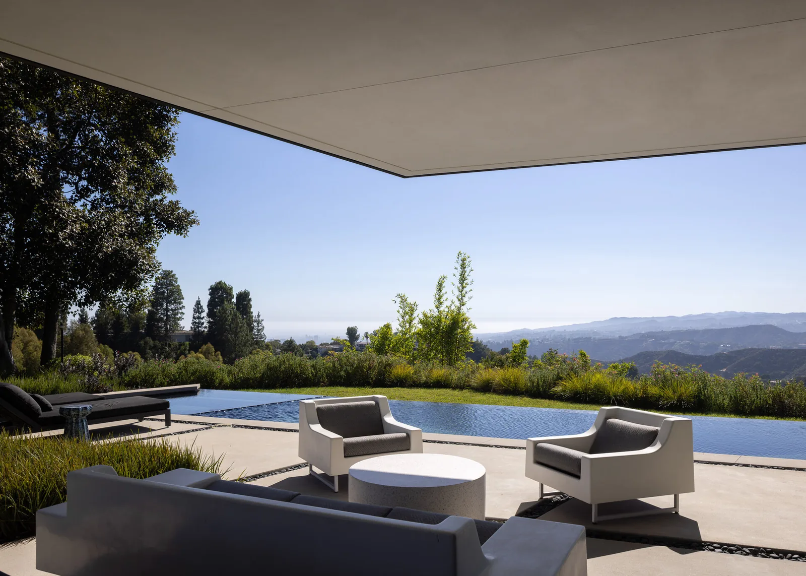 Patio seating area beside an infinity pool with views of landscaped greenery and distant mountains.