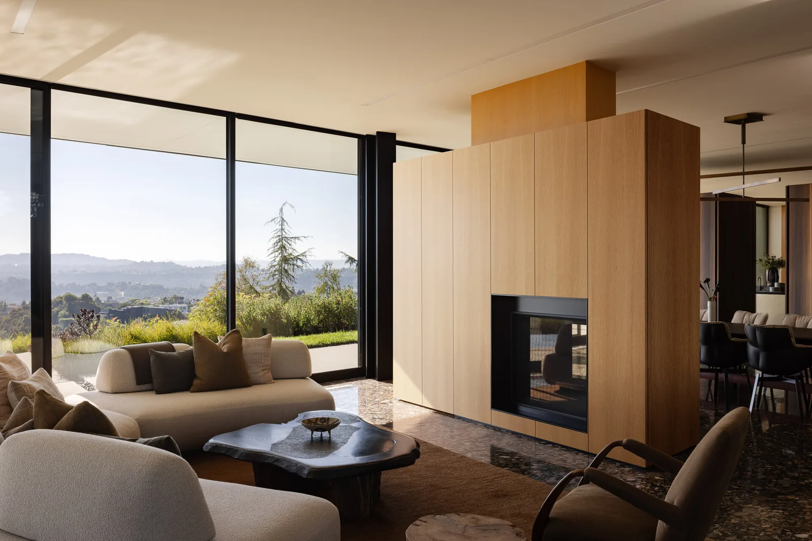 Living area with large glass windows and a central wood-paneled fireplace, overlooking landscaped garden and distant mountains.