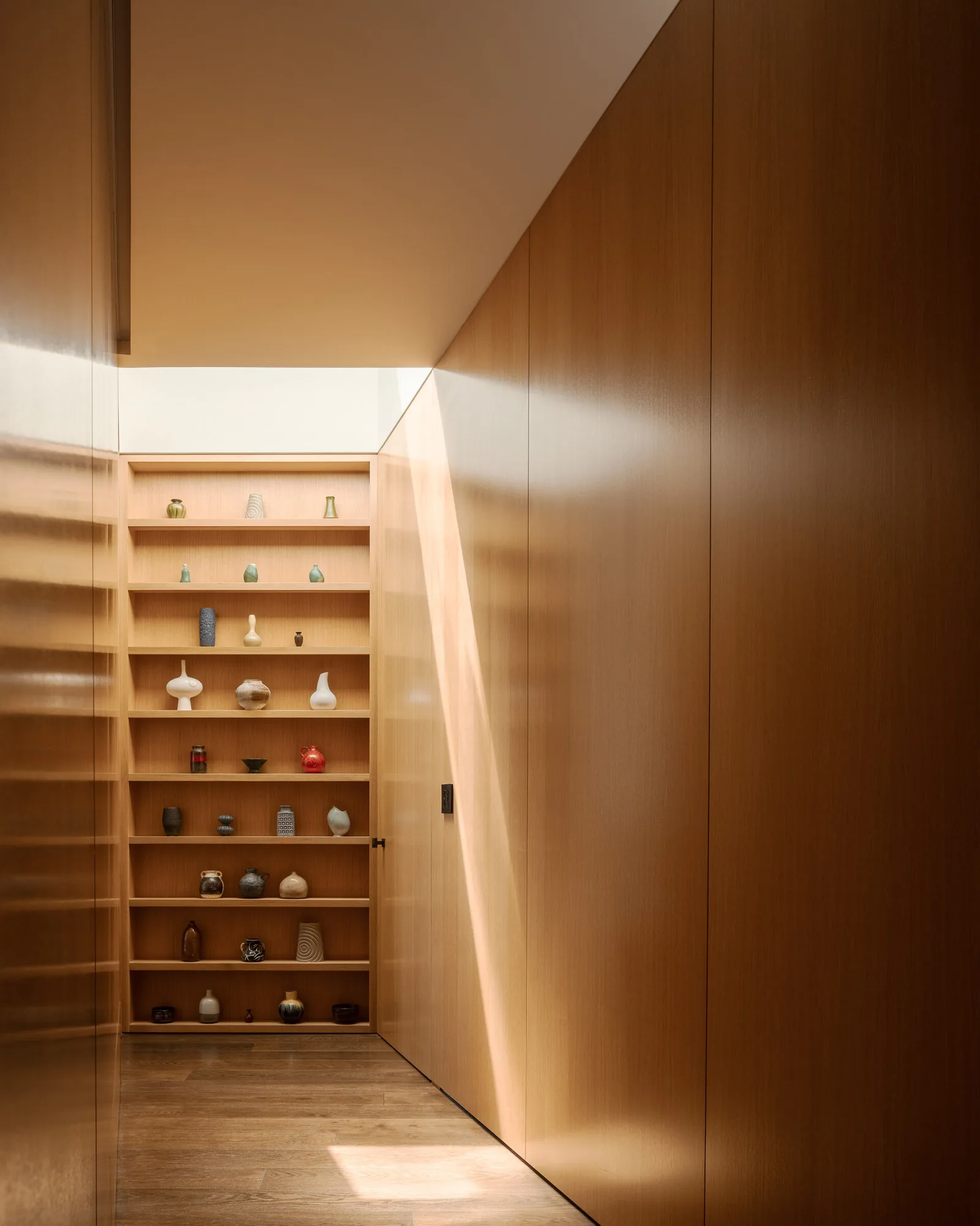 A hallway features built-in wood shelves displaying pottery and light wood panel walls illuminated by skylight.