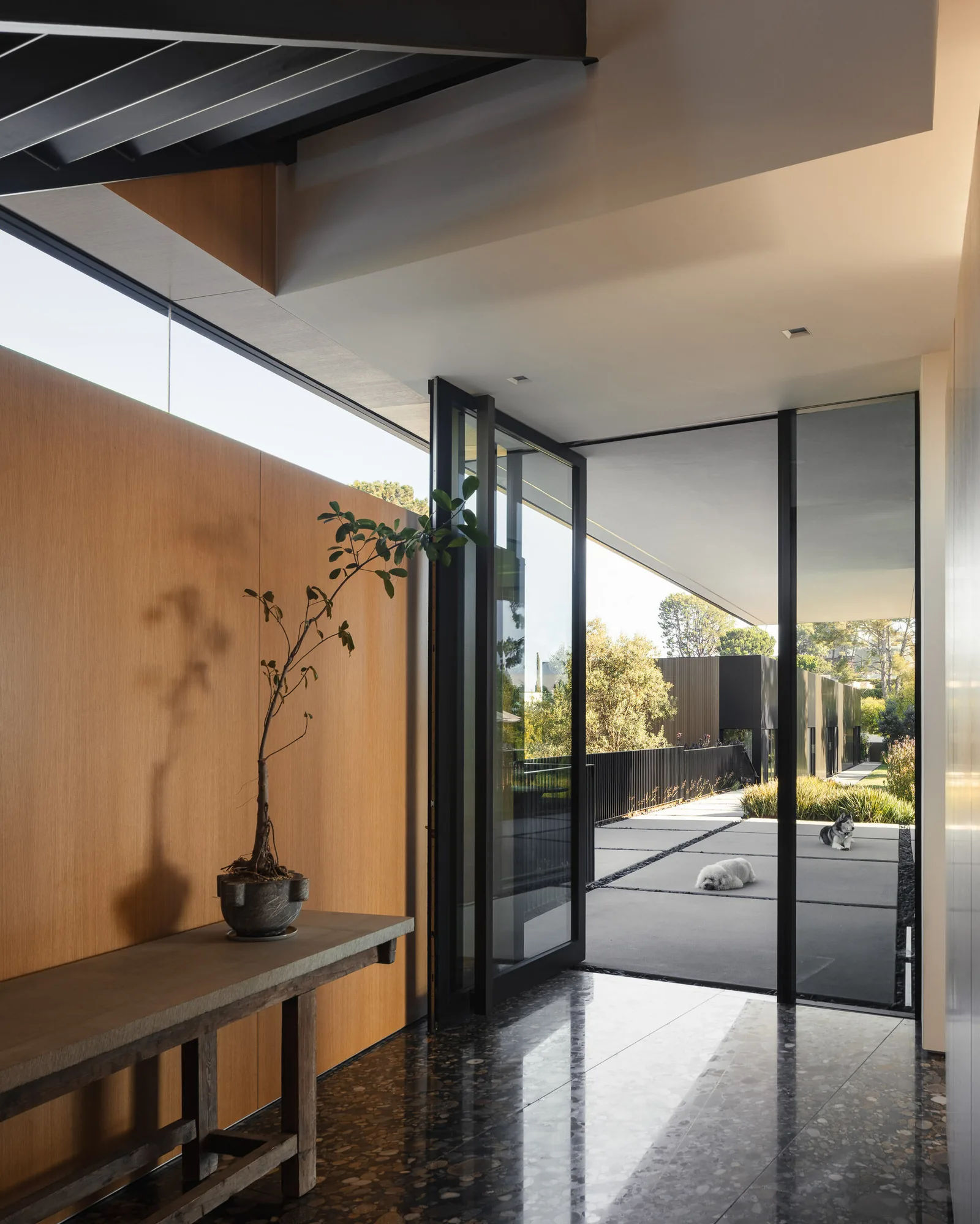 Entry area with terrazzo floor, wood-paneled wall, and black-framed glass door opening to outdoor pathway and garden.