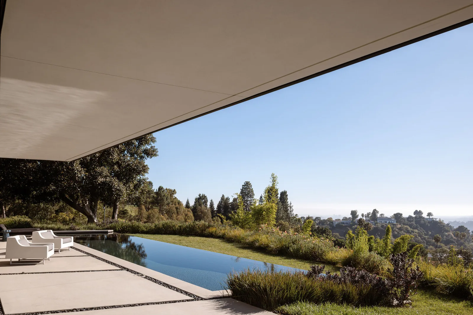 The patio features white outdoor seating, an infinity pool, and a large overhanging roof blending into the landscaped garden view.