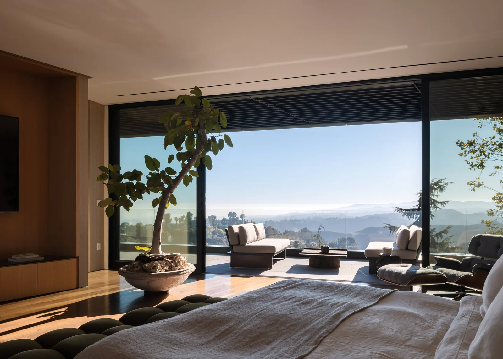Bedroom with a potted tree indoors and outdoor seating overlooking distant mountains through floor-to-ceiling glass doors.