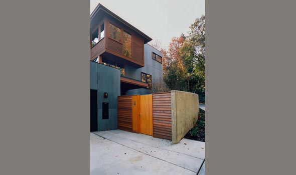 Modern multi-story residential building with wood and concrete facade, featuring horizontal wooden slats, large windows, and an entry gate.