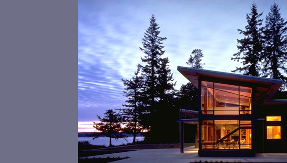 Evening view of the Bruckner Residence showing a two-story house with extensive floor-to-ceiling glass windows and a sloped roof, set among,
