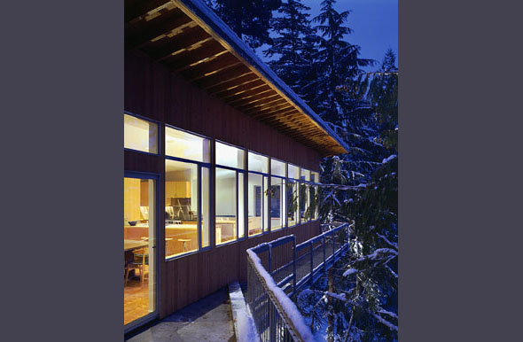 Evening view of a wood-sided house exterior with a metal railing balcony and snow on trees nearby.