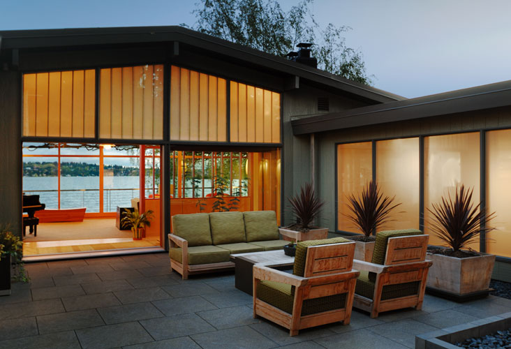 Outdoor patio seating area with wooden frame furniture and a view into an interior room with large glass windows overlooking water.
