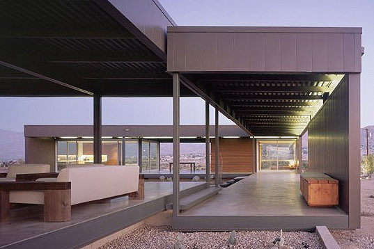 Exterior view of a modern desert house with a covered patio, steel frame, and large glass windows showing interior and cityscape beyond.